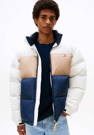 Un jeune homme porte une doudoune à blocs de couleurs blanc, beige et bleu marine, par-dessus un pull bleu marine et un jean bleu clair, sur un fond blanc uni.