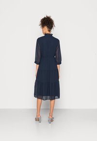 Námořnické modré šaty s volánkovým límcem a tříčtvrtečními rukávy, vyrobené z lehkého materiálu. Doplněné kostkovanými sandály na blokovém podpatku.