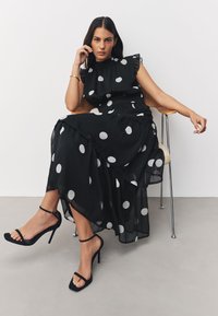 Vestito nero a pois con maniche arricciate, gonna a strati e tessuto trasparente; abbinato a tacchi alti neri con cinturini su un pavimento neutro.