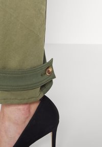 Olijfgroene broek met opgerolde mouwen, voorzien van een beige knoop. Zwarte hoge hak schoenen met een spitse neus en een gestructureerde suède afwerking.
