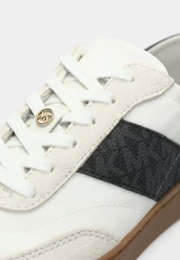 Sapatilha branca com detalhes em camurça bege e uma faixa lateral preta com padrão. Apresenta fecho de atacadores e um pequeno emblema dourado perto dos atacadores.