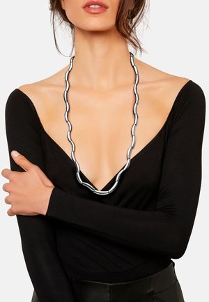 Collier en métal argenté présentant des maillons allongés et courbés avec une finition lisse, porté sur un haut noir à manches longues avec un décolleté en V profond.