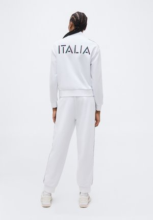 Osoba z warkoczykami, ubrana w biały dres z napisem "ITALIA" na plecach, stojąca na tle jednolitego białego tła.