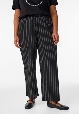 Osoba v čiernych širokých nohaviciach s bielymi zvislými pruhmi, v čiernom tričku a čiernych topánkach Mary Jane, stojaca na bielom pozadí.