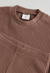 Braunes Baby-Outfit mit Waffelstruktur, Rundhalsausschnitt und Druckknopfdetails. Weicher Stoff mit gerippten Bündchen und lockerem Schnitt.