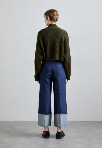 Person wearing dark green ribbed sweater and wide-leg cuffed blue jeans, standing barefoot on a gray floor against a plain wall.