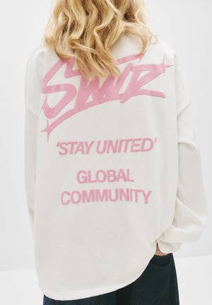Hvid oversized sweatshirt med lyserød grafisk tekst på ryggen: "Swiz," "'Stay United'," og "Global Community." Blødt stof, afslappet pasform.