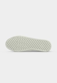 Semelle de chaussure blanche avec un motif en relief de losanges et d’étoiles, vue du dessous sur un fond gris clair uni.
