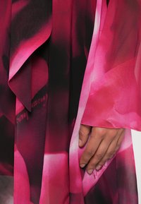 Mão repousada em tecido fluido com padrões florais abstratos em tons de rosa e vermelho escuro e textura delicada e translúcida.