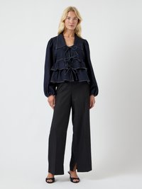 Blusa de ganga com grande colar, três camadas de franzido e detalhe em laço. Costura castanha contrasta com o tecido escuro. Combinada com calças largas pretas.