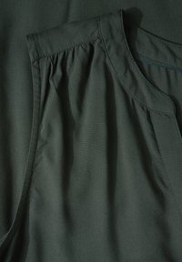 Tmavě zelený textilní top s jemnou texturou, s nařasenými detaily na rameni a hladkým lemováním kolem výstřihu.