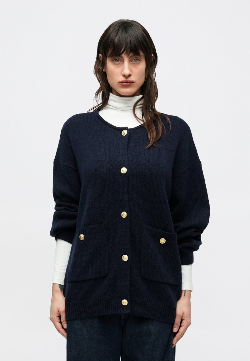 Cárdigan azul marino de tela texturizada, con grandes botones dorados y dos bolsillos delanteros. Corte holgado con hombros caídos.