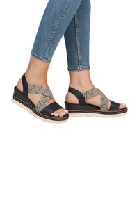 Black wedge sandals with woven geometric-patterned straps, beige sole, and open toe. Features a cushioned footbed and adjustable ankle strap.