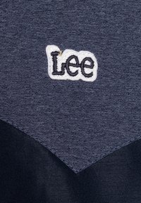 Denimová látka v tmavě modré barvě, s bílou vyšitou značkou "Lee" a kontrastní tmavou látkovou částí na spodní straně.