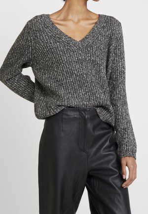 Žena v šedém texturovaném svetr s výstřihem do V, zastrčeném do černých kožených kalhot s vysokým pasem, stojící s jednou rukou pokrčenou.