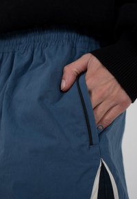 Hand teilweise in der Reißverschlusstasche einer blauen Hose mit elastischem Bund, getragen zu einem schwarzen gerippten Pullover.