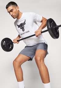 Homme portant un t-shirt blanc Project Rock et un short gris, soulevant une barre avec des poids noirs, concentré sur son entraînement.