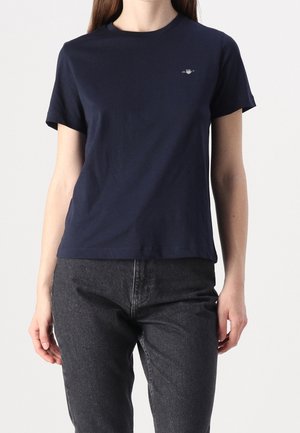 Femme portant un t-shirt uni bleu marine à manches courtes et un jean taille haute gris foncé sur un fond clair.
