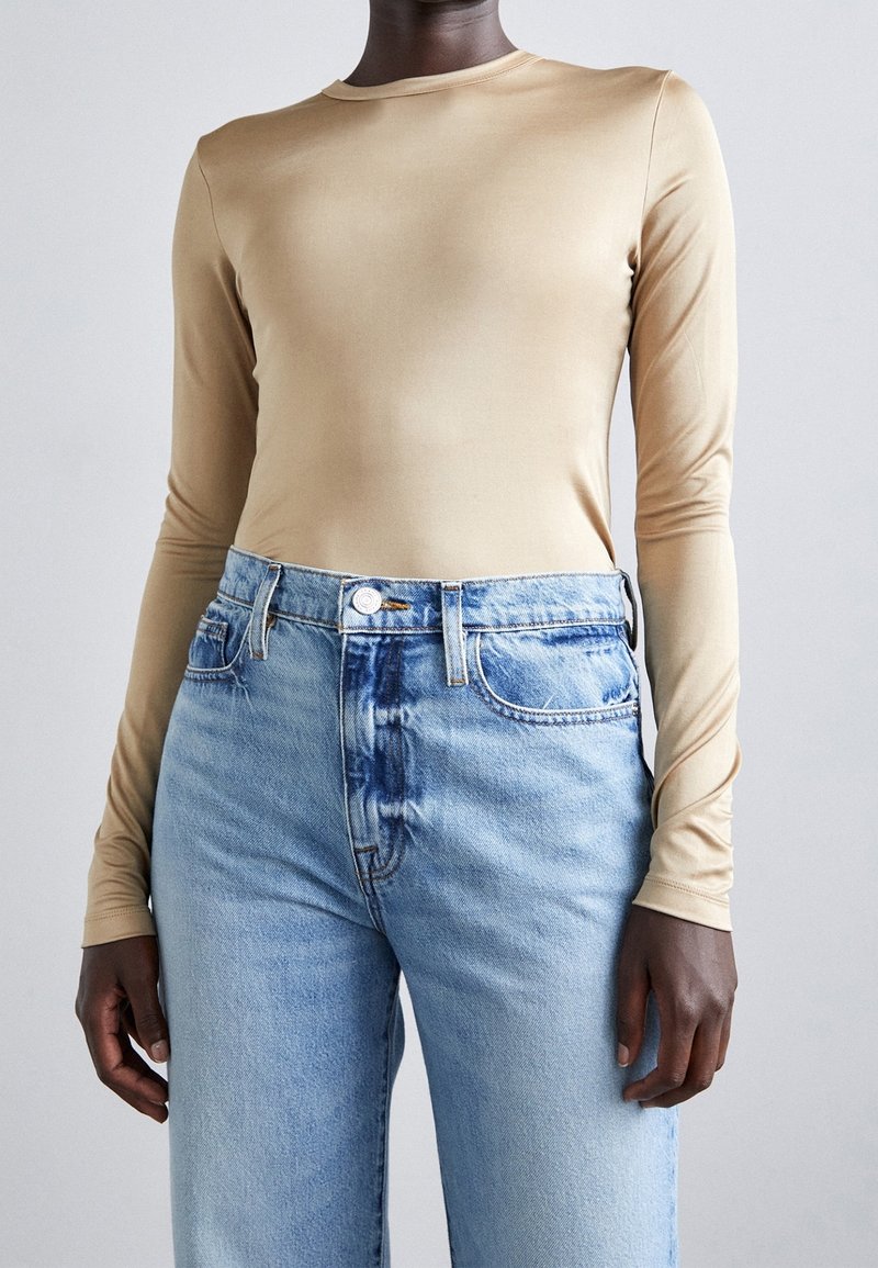 Beige Langarmoberteil aus dehnbarem Stoff, kombiniert mit hellblauen, hoch taillierten Jeans mit verwaschenem Textur und sichtbaren Nähten.