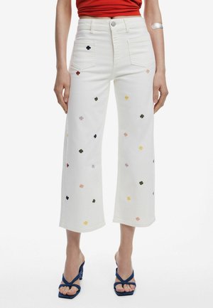 Hvide, vidde, korte bukser med flerfarvede broderede blomster, forlommer og knaplukning, båret med blå sandaletter med hæl.