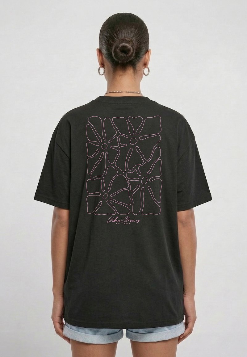 Femme avec les cheveux en chignon portant un t-shirt noir oversize avec un motif floral abstrait rose en traits linéaires et le texte « Urban Classics » dans le dos.