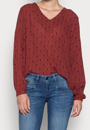Femme portant un haut à manches longues couleur rouille avec un motif géométrique et un jean en denim bleu, se tenant devant un fond clair.