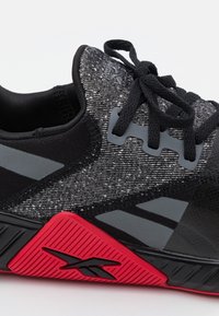 Close-up van een zwart en grijs sportshoe met een gestructureerde bovenkant, zwarte veters en een rode zool met een zwart logo.