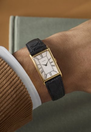 Poignet portant une montre Timex rectangulaire en or avec cadran blanc, chiffres romains noirs, et bracelet en cuir noir sur une manche de veste en velours côtelé marron.