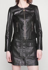 Giacca in pelle nera con chiusura a zip e due tasche frontali, abbinata a una gonna in pelle coordinata con bottoni a pressione e tasche laterali.