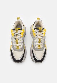 Tömeg sneaker fehér, szürke és fekete színben sárga részletekkel. Jellemzői a hálós és szintetikus anyagok, kerek orr és párnázott gallér.