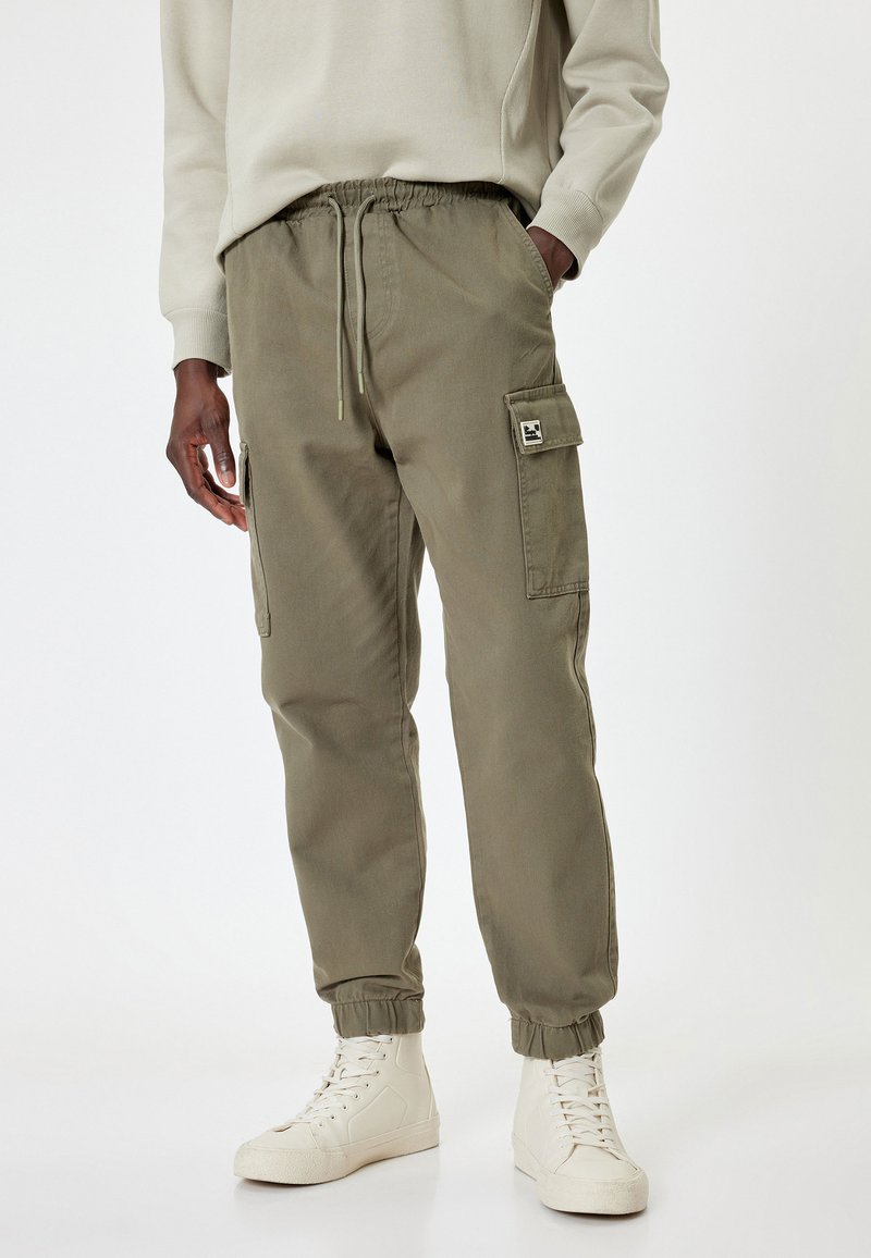 Cargo broek in olijfgroen met een elastische tailleband, trekkoord, zijzakken en een patchlogo op de bovenbeen. Zacht, stevig materiaal.