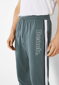 Türkise Jogginghose mit einem Kordelzug, weißen Seitenstreifen und großen "Bench."-Text in Weiß, aus einem weichen Stoff gefertigt.