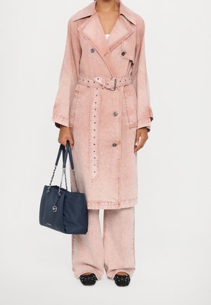 Trench-coat en denim rose clair avec un avant croisé, une ceinture et des clous. Associé à un sac bleu marine et des chaussures noires ornées de clous.