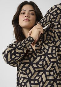 Beige Bluse mit langen Ärmeln und einem schwarzen geometrischen Muster. Sie verfügt über einen V-Ausschnitt und elastische Bündchen für zusätzlichen Komfort und eine optimale Passform.