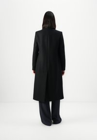 Cappotto lungo nero con silhouette strutturata, con spacchetto singolo sul retro e texture liscia. Indossato sopra pantaloni scuri, design minimalista.
