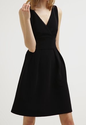 Mouwloze zwarte jurk tot op de knie met V-hals en getailleerde taille, gedragen door een vrouw tegen een effen lichte achtergrond.