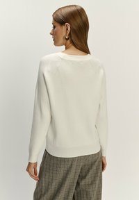 Biały sweter w prążki z okrągłym dekoltem i długimi rękawami, w połączeniu z wzorzystymi szarymi spodniami. Tekstura wydaje się miękka i przytulna.