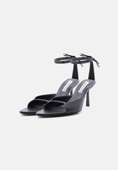 Ζευγάρι μαύρα πέδιλα με ψηλό τακούνι, ανοιχτά στα δάχτυλα, με λουράκια στον αστράγαλο και τετράγωνη μύτη σε λευκό φόντο.