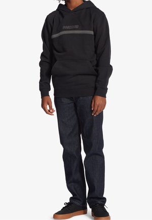 Personne portant un sweat à capuche noir DC Shoes, un jean bleu foncé et des baskets noires à semelles marron, debout devant un fond blanc uni.