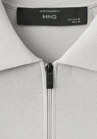 Col montant de chemise en maille gris clair avec une fermeture éclair noire et une étiquette noire indiquant "PERFORMANCE MNG EU-USA M MEX M" à l'intérieur.