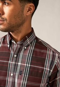 Camisa a cuadros burdeos con cuello abotonado, que presenta líneas blancas y negras, doble botón en el cuello y un pequeño logo en el bolsillo del pecho.