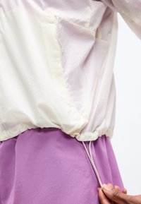 Χέρι που τραβάει το κορδόνι στη μέση ενός λευκού αδιάβροχου μπουφάν πάνω από μοβ παντελόνια σε ανοιχτό φόντο.