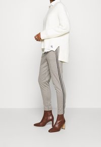 Pull en maille crème oversize superposé sur une chemise blanche à col, associé à un pantalon à carreaux avec des bandes latérales et des bottines à talons marron.