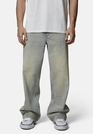 Personne portant un jean bleu délavé et ample à jambes larges et des baskets blanches, debout devant un fond uni.