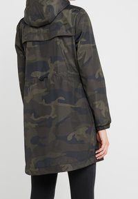 Achteraanzicht van een persoon die een donkergroene en zwarte camouflage parka met capuchon draagt, met elastische manchetten en een verstelbaar taillekoord.