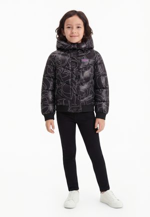 Un jeune enfant se tient debout, portant une doudoune noire à capuche avec des lettres blanches abstraites, un pantalon noir et des baskets blanches sur un fond uni.