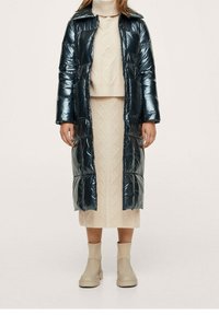 Skinnende mørkeblå lang dynefrakke med stor krave og to forlommer, båret over en beige strikket nederdel og højhalset trøje, kombineret med beige ankelstøvler.