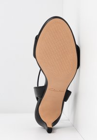 Sandale à talon haut noire montrée de la semelle avec un dessous texturé beige et le logo de la marque Clarks, positionnée contre un fond blanc d’angle.
