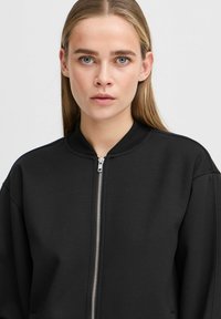 Svart zip-up bomberjacka med ribbad krage och textilstruktur. Har en silverfärgad dragkedja och en avslappnad passform. Minimalistisk design.