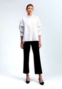 Camicia bianca con bottoni e spalle scese, abbinata a pantaloni neri a gamba larga e décolleté neri a punta. Tessuto liscio, design minimale.